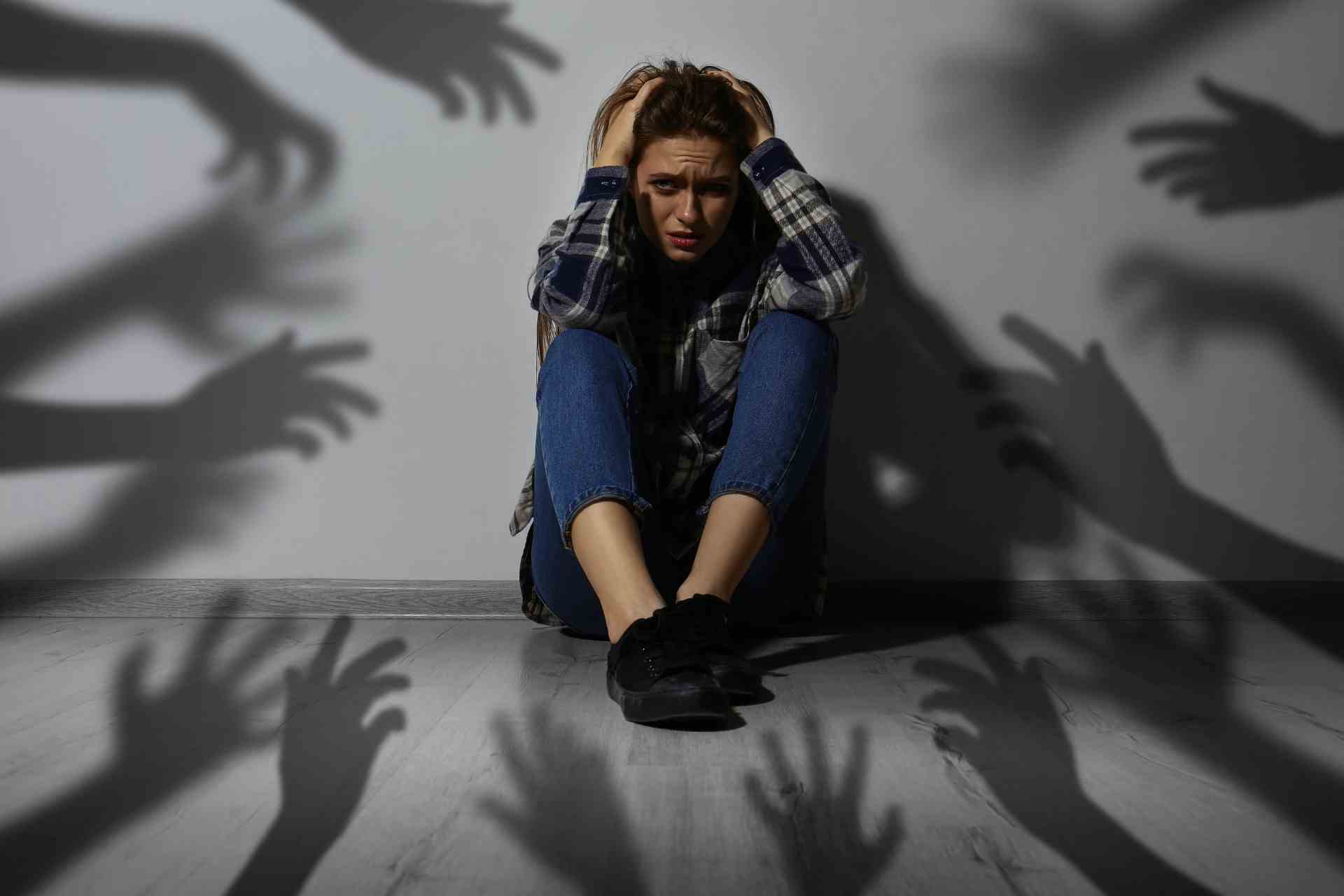 Woman sitting on the floor in distress with shadowy hands around her, representing social anxiety and overwhelming fear