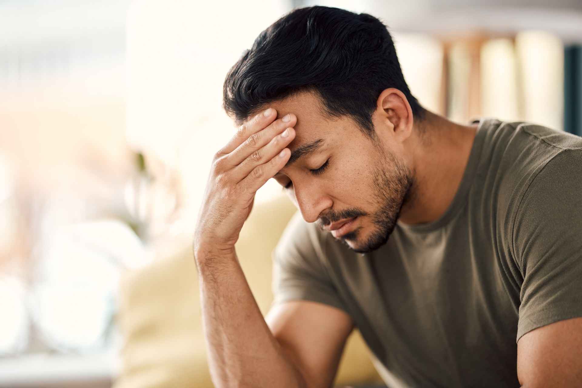 Man sitting with his eyes closed and one hand pressed against his forehead, appearing stressed and overwhelmed.