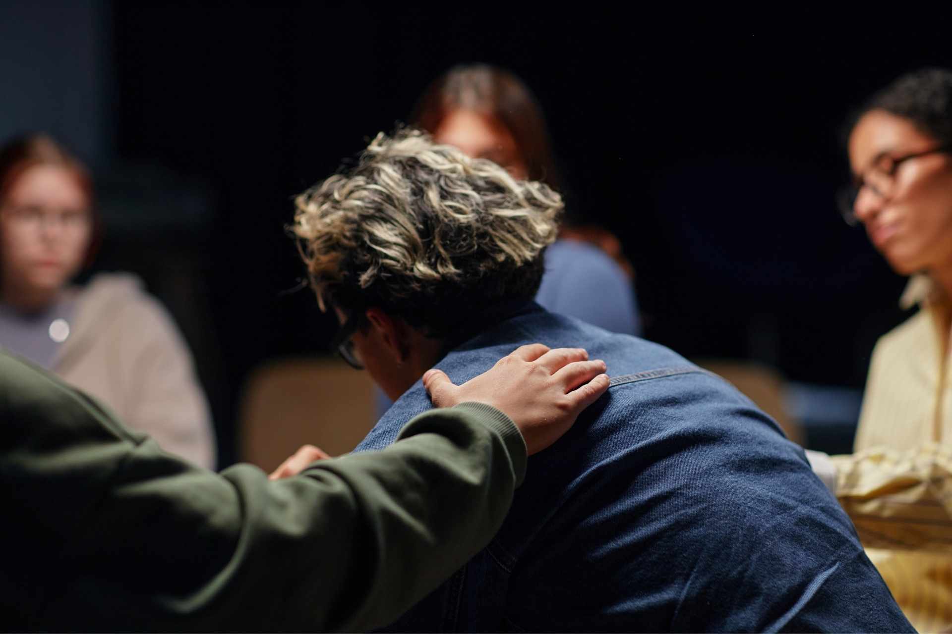 Person placing a comforting hand on another person's shoulder during a seated group conversation.
