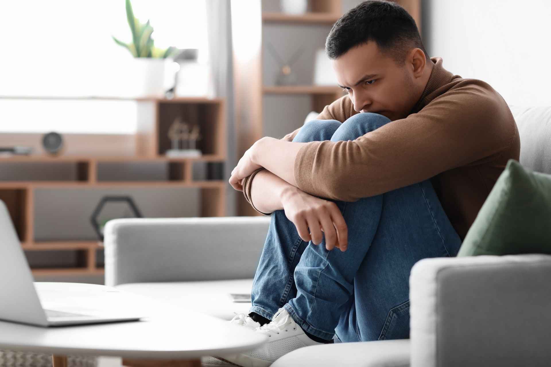 Young man sitting alone on a couch with his knees drawn toward his chest, looking worried and distressed.